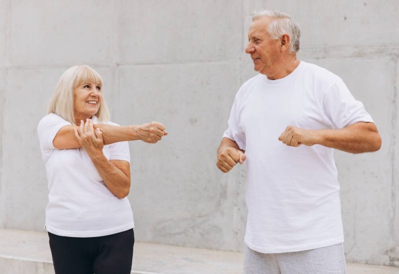 Senior couple exercising outdoors, performing arm stretches as part of their fitness routine. They are wearing casual athletic wear and sharing a moment of healthy activity and companionship.